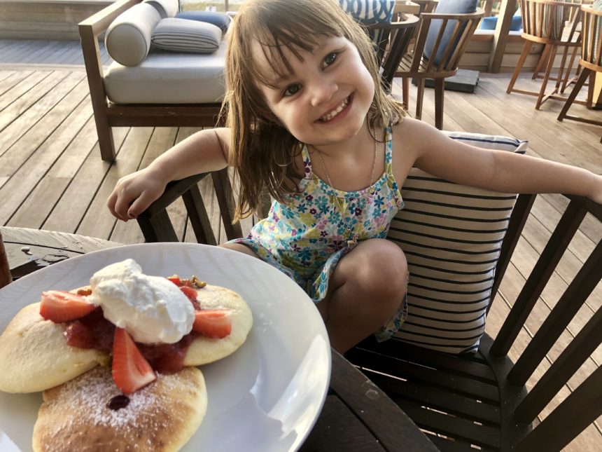 a girl sitting at a table with a plate of pancakes and strawberries